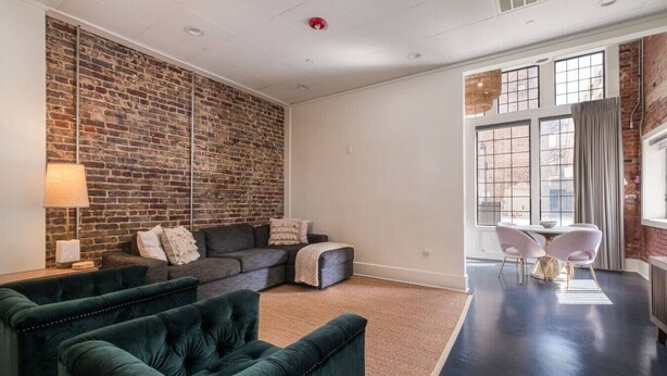 Ein modernes Wohnzimmer mit Backsteinwand, einem Ecksofa, einem grünen Sessel und einem Essbereich mit weißen Stühlen.