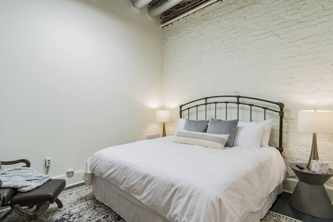 Een slaapkamer met een wit bed, hoofdbord, twee bedlampen, een kleine tafel, een stoel en een witte bakstenen muur.
