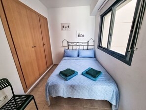 A bedroom with a bed, a wooden wardrobe, a window, and a small wall-mounted shelf with decorative items.