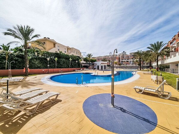 A swimming pool with sun loungers and a water fountain, surrounded by palm trees and buildings.