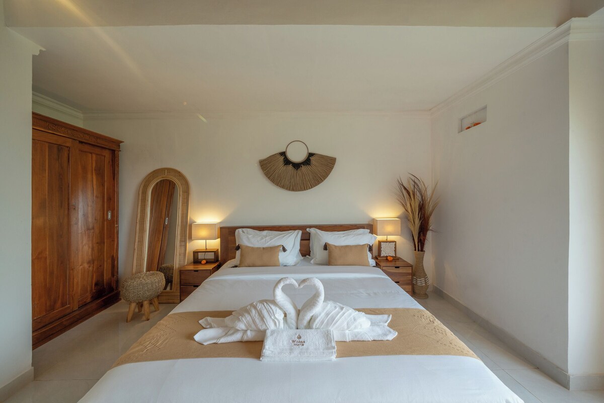 A hotel room with a large bed, wooden wardrobe, a mirror, and a wall-mounted fan.