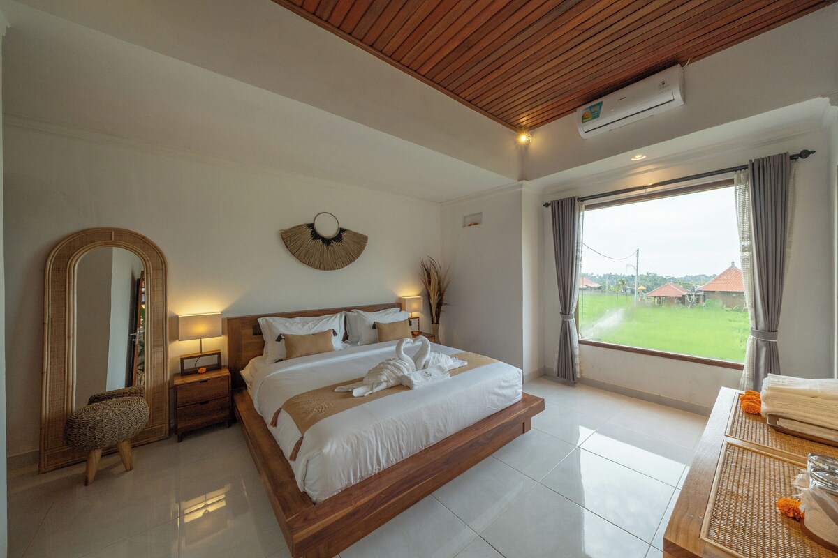 A spacious bedroom with a large bed, a wooden ceiling, a large window with a view of the outdoors, and a bedside table with a lamp.