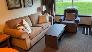 Ein Wohnzimmer mit einem braunen Sofa, einem Holztisch, einem grauen Sessel und einem großen Fenster mit Blick auf eine grüne Rasenfläche.