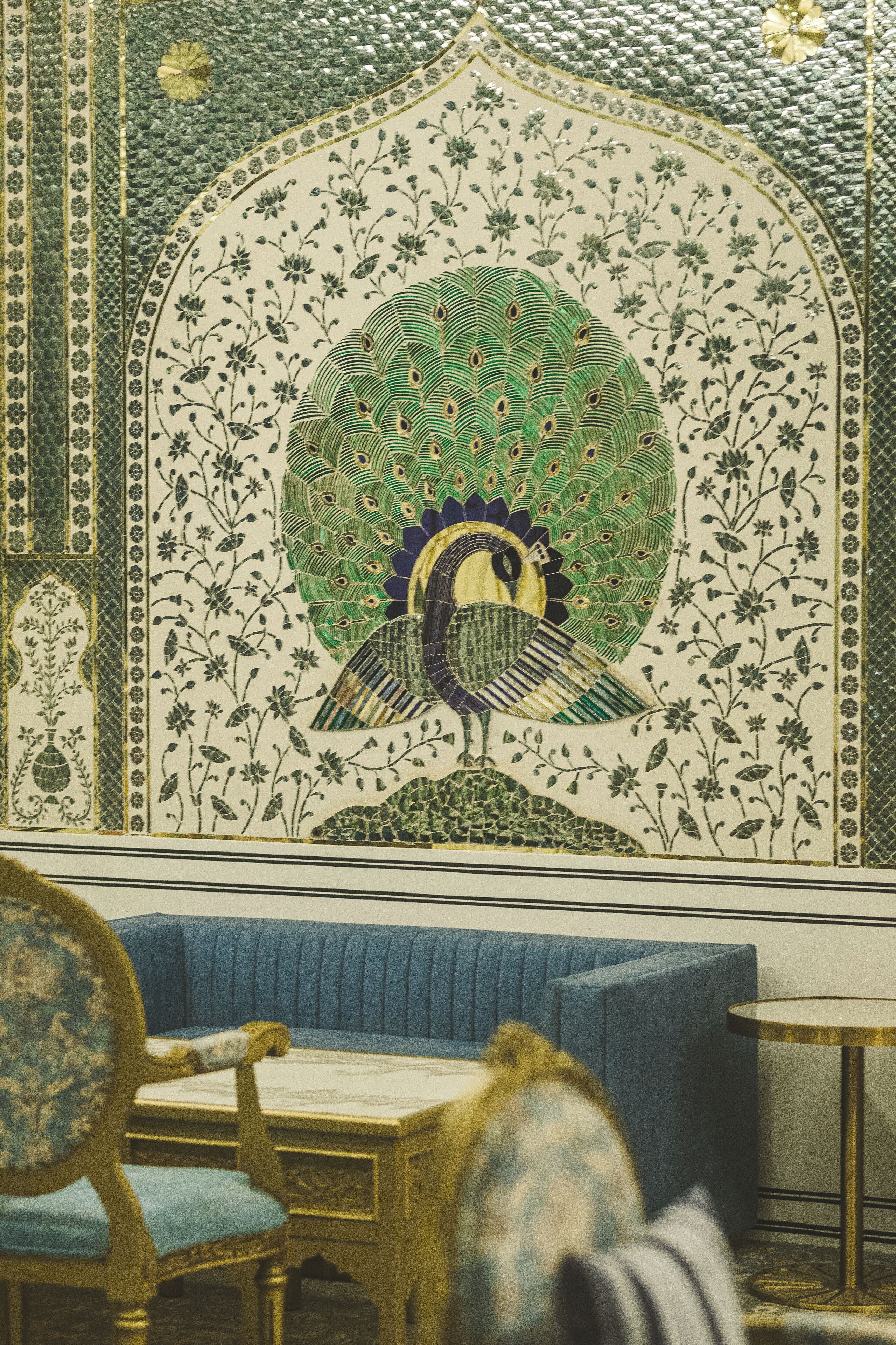 Una habitación con un mosaico de pavo real en la pared, un sofá tapizado en azul y una mesa redonda con base dorada.