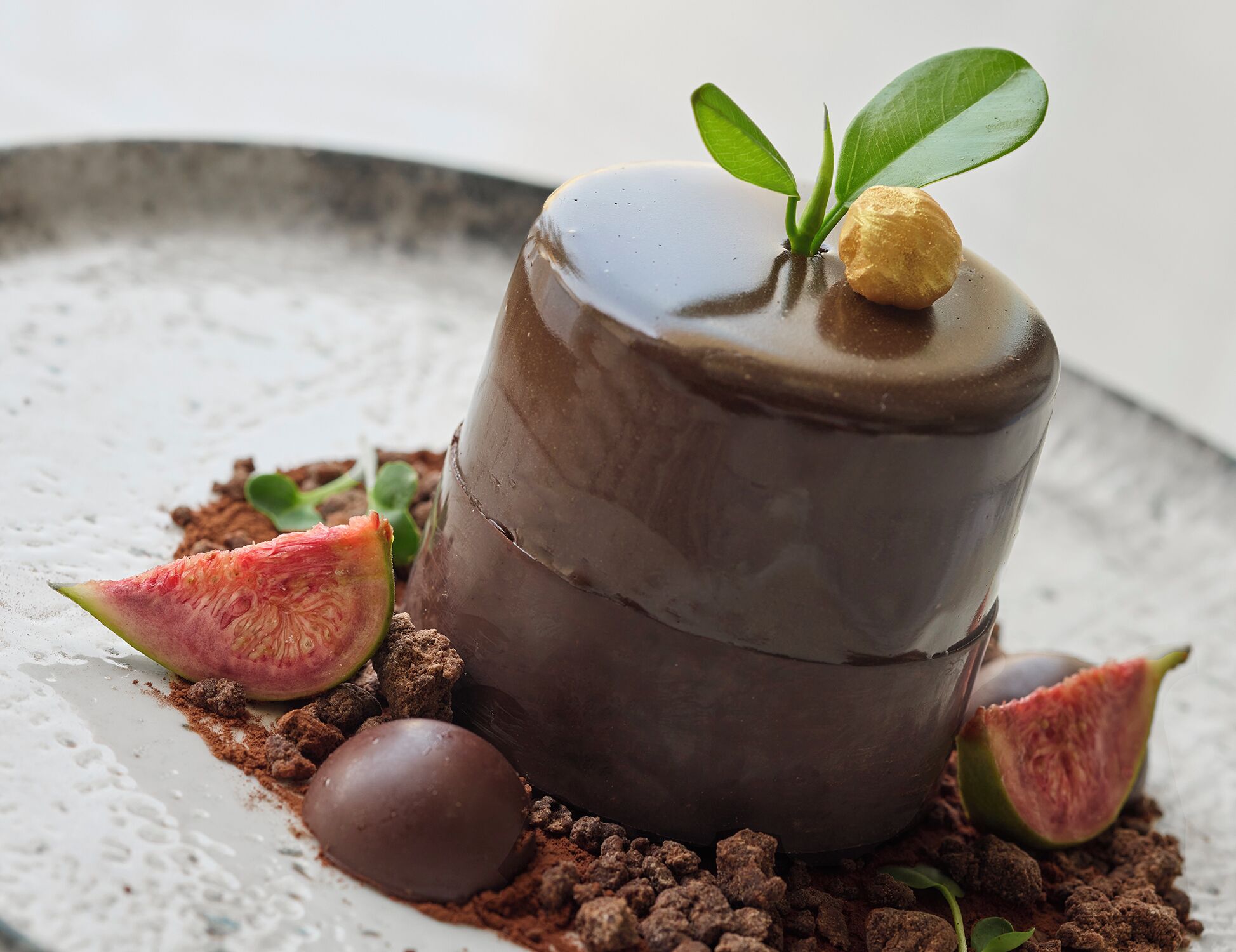 Sobremesa de chocolate com cobertura brilhante, acompanhada de uma folha verde e uma fatia de fruta.