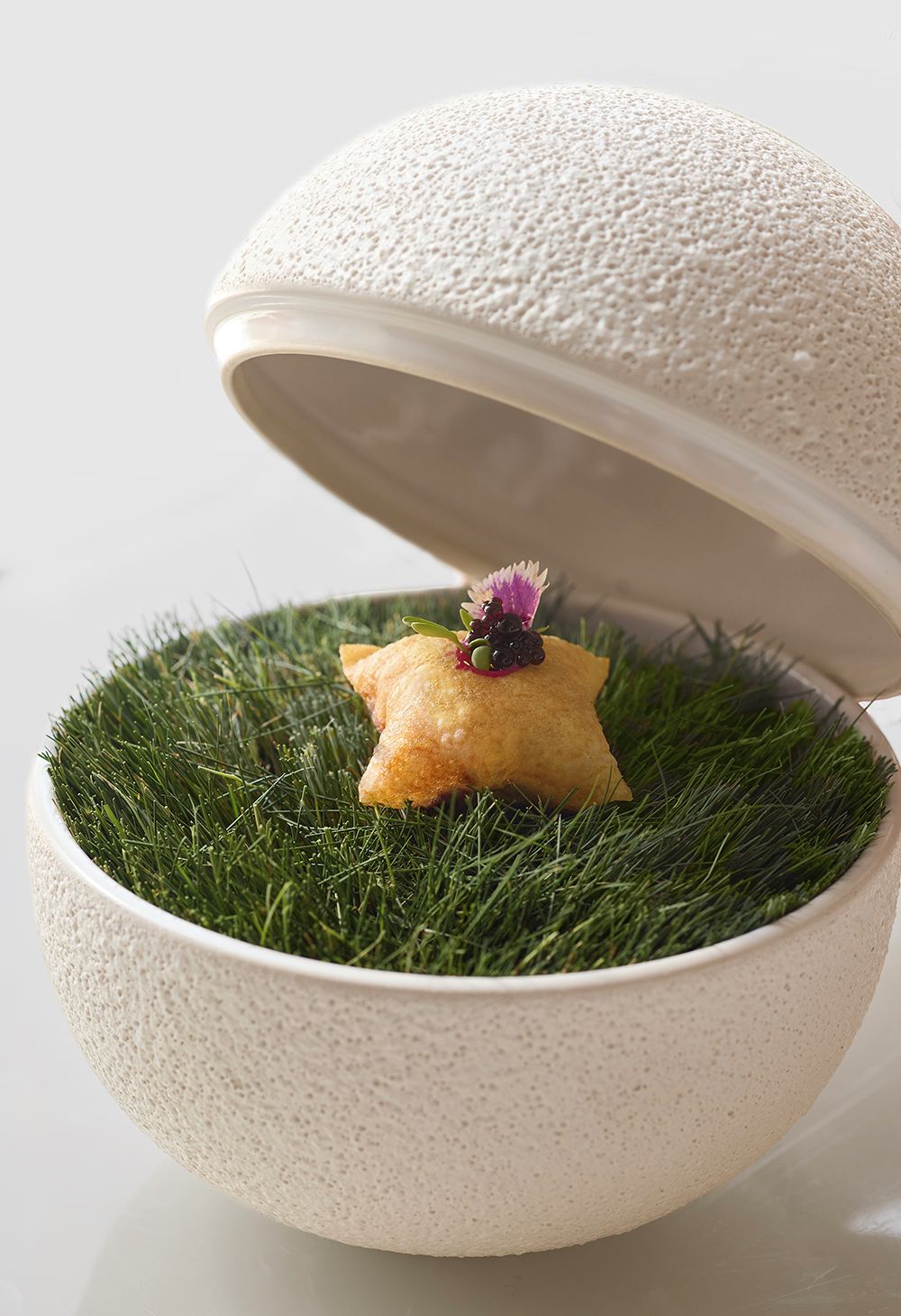 Un bol con superficie texturizada y tapa, que contiene un pastel con forma de estrella sobre un lecho de hierba verde, con una flor decorativa encima.