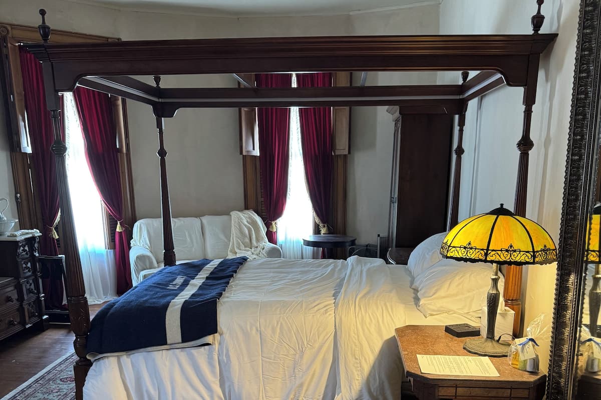Una cama con dosel, un tapete con estampado floral, una mesita de noche con lámpara y un espejo en la pared.