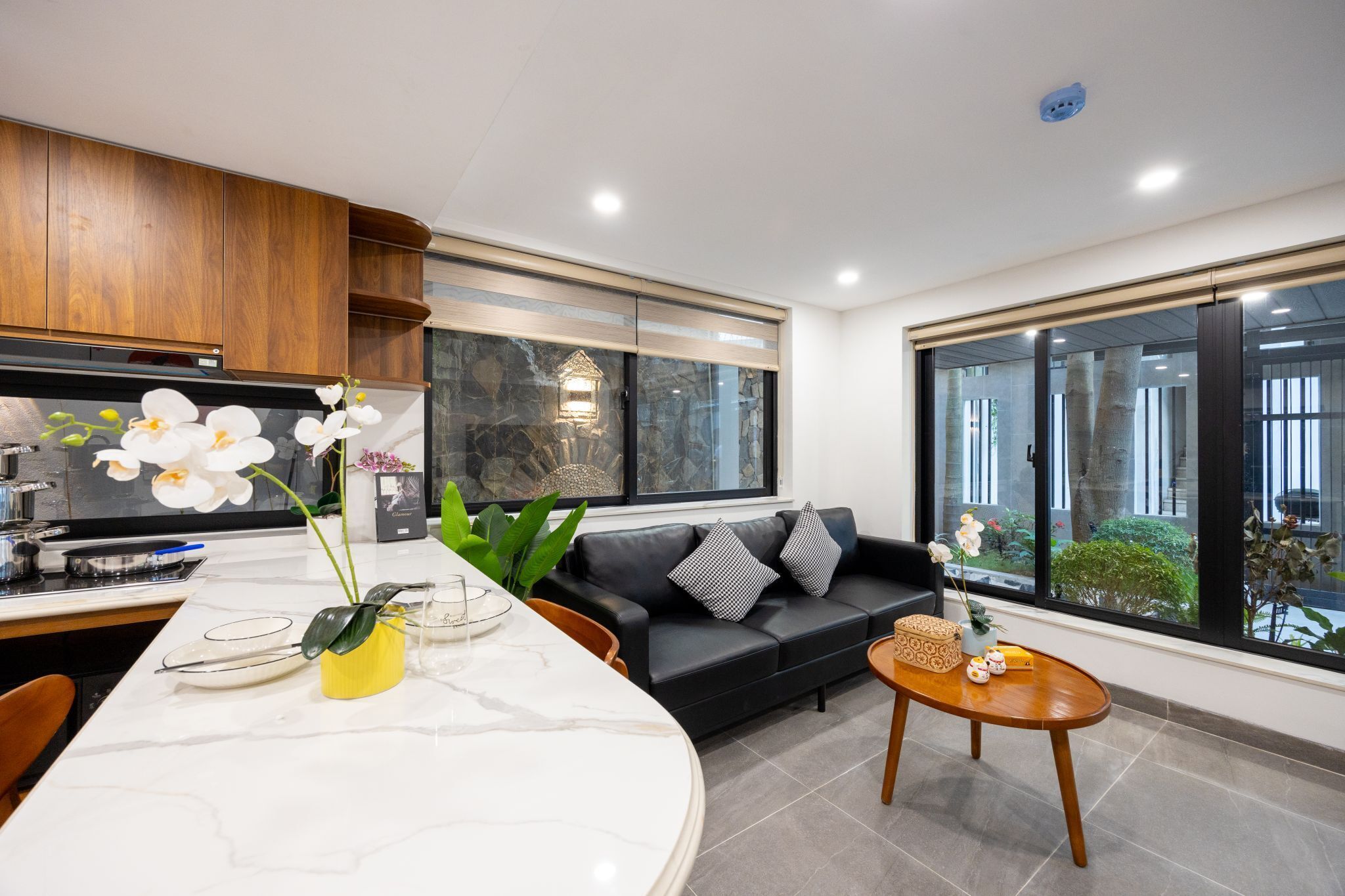 A modern living room with a black sofa, a marble dining table, and a large window offering a view of the outdoors.