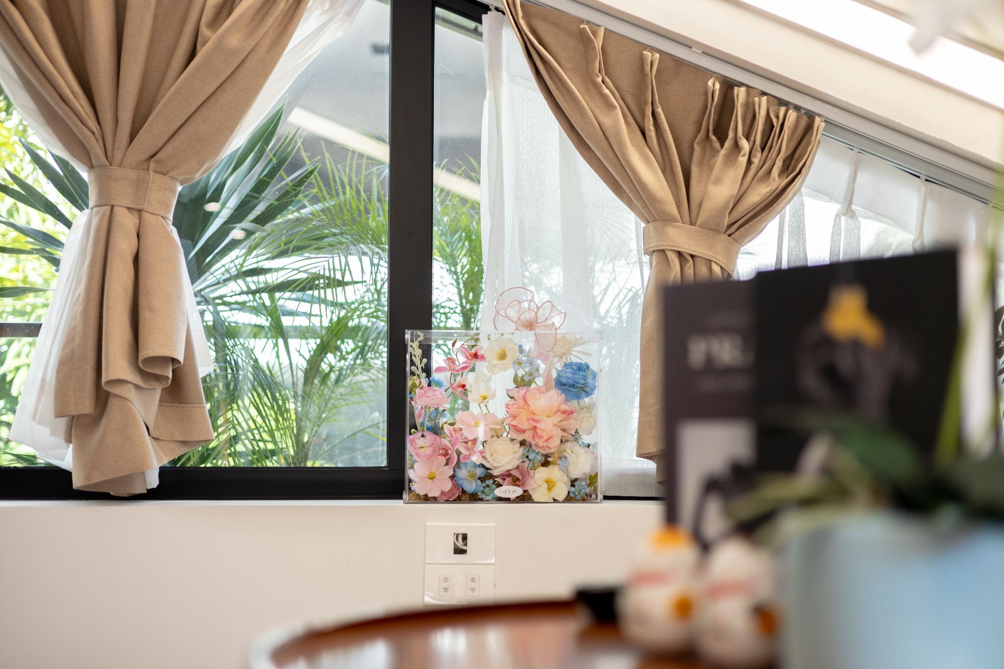 A window with beige curtains, a floral arrangement, and a framed picture on a table.