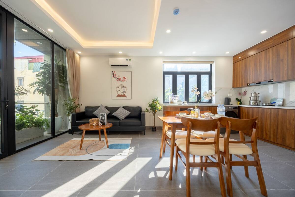A modern living room with a sofa, coffee table, and dining area. There is a kitchen with wooden cabinets and a window.