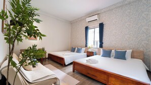 Ein Hotelzimmer mit zwei Einzelbetten, einem großen Bett, Holzboden, einer Pflanze und einem Fenster mit Vorhängen.