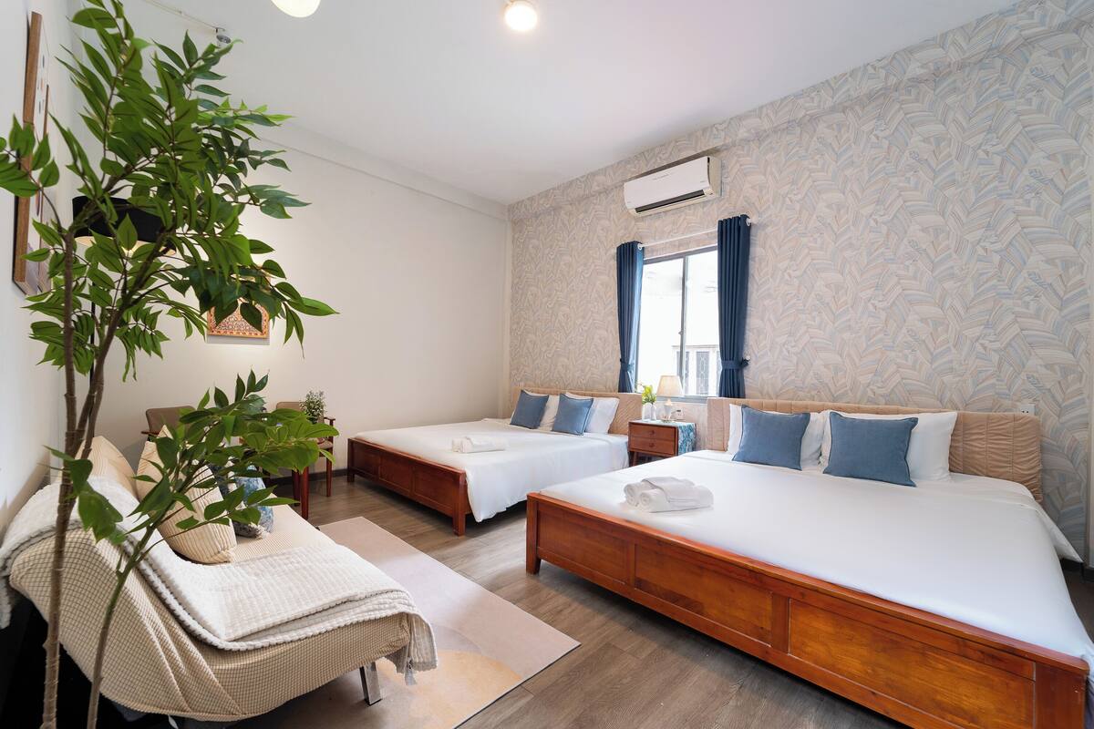 A hotel room with two single beds, a large bed, a wooden floor, a plant, and a window with curtains.