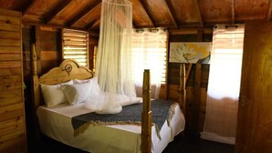 A wooden cabin bedroom with a bed, mosquito net, and a painting on the wall. - Take it easy at our eco friendly beach cabins where life meets nature funfilled  (Westmoreland)