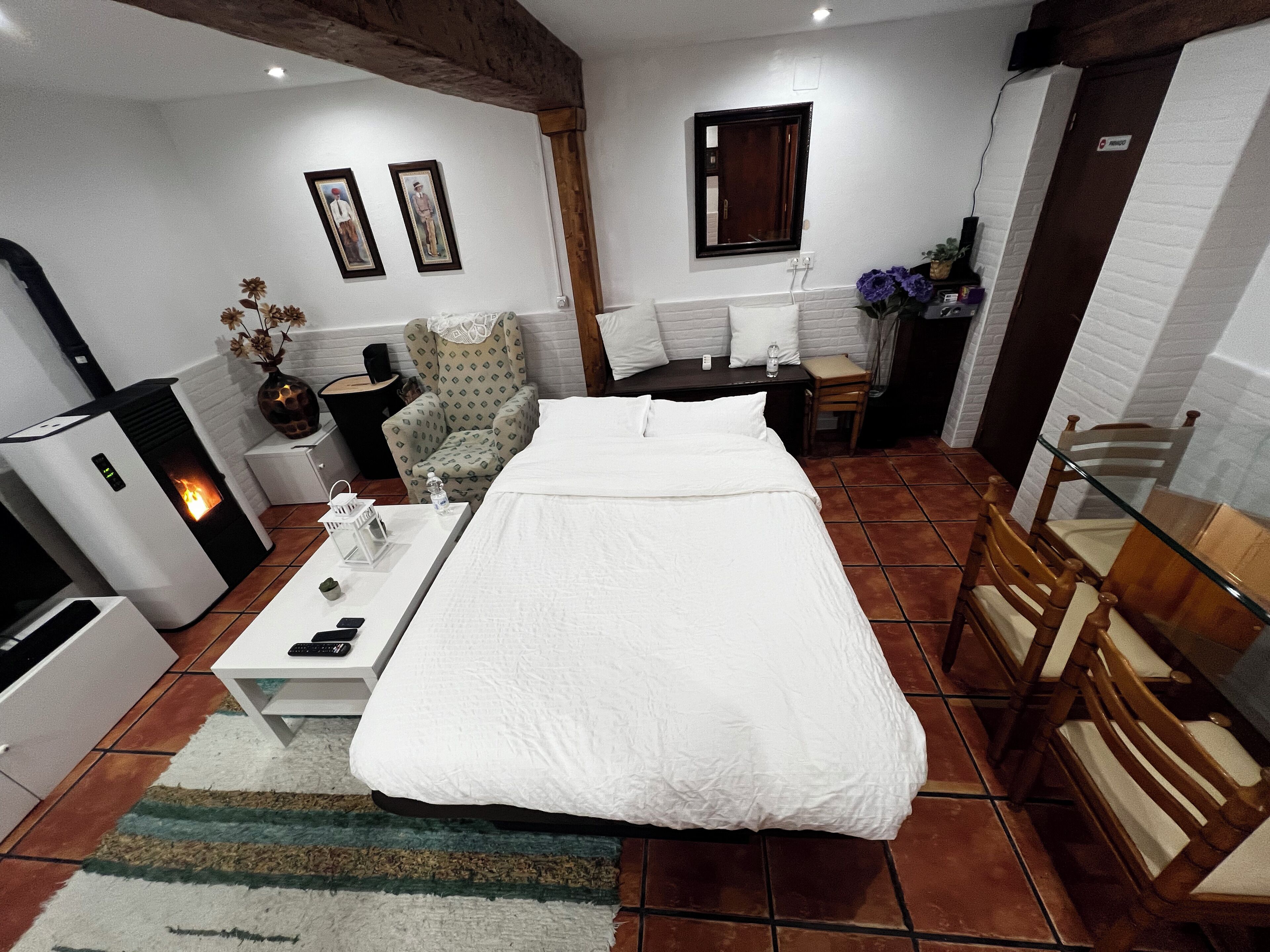 A hotel room with a bed, a desk with a chair, a small table with a vase, a heater, and a mirror on the wall.