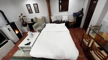 A hotel room with a bed, a desk with a chair, a small table with a vase, a heater, and a mirror on the wall.