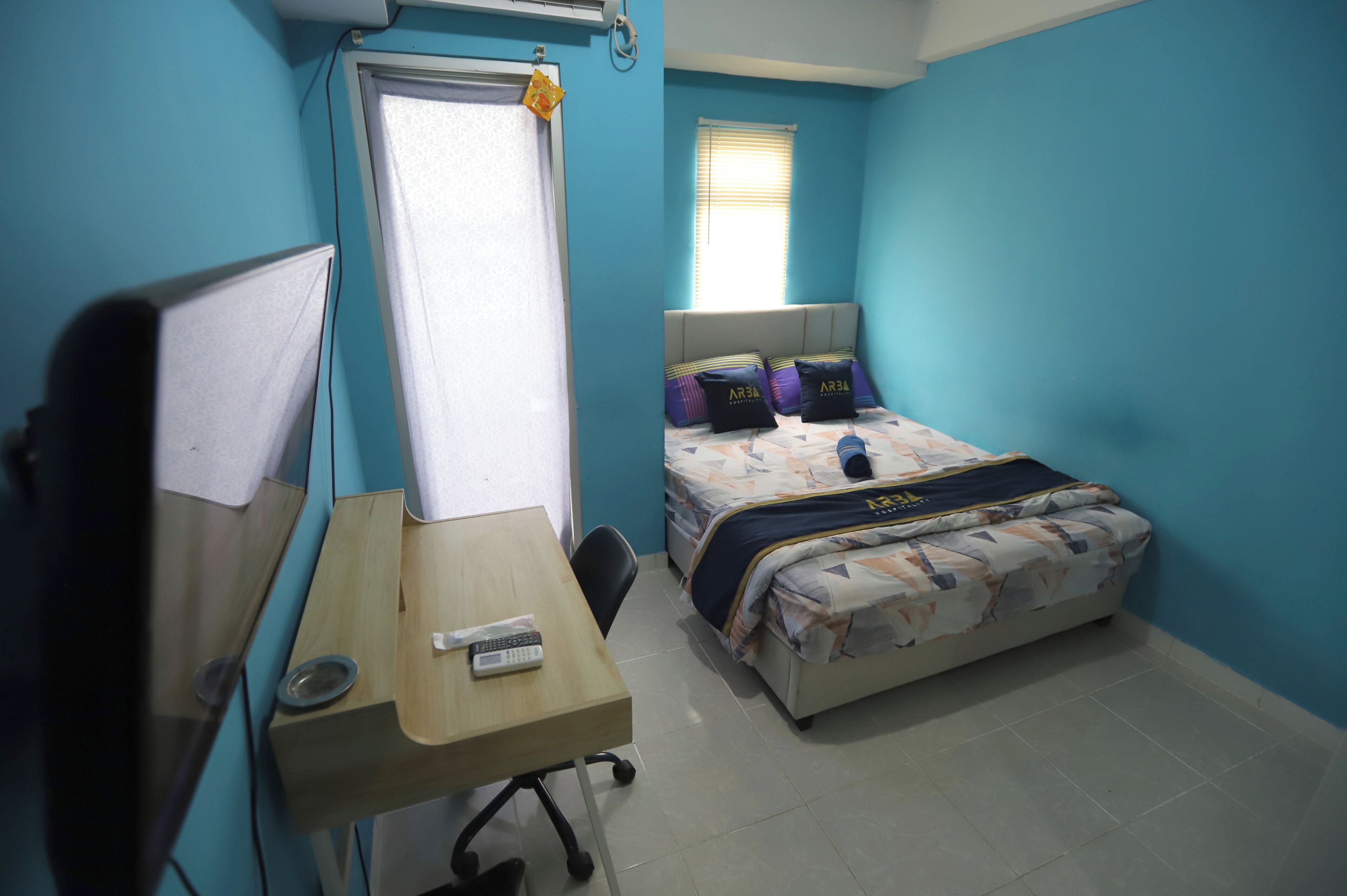 A bedroom with a bed, a desk with a chair, a window with blinds, and a wall-mounted air conditioner.