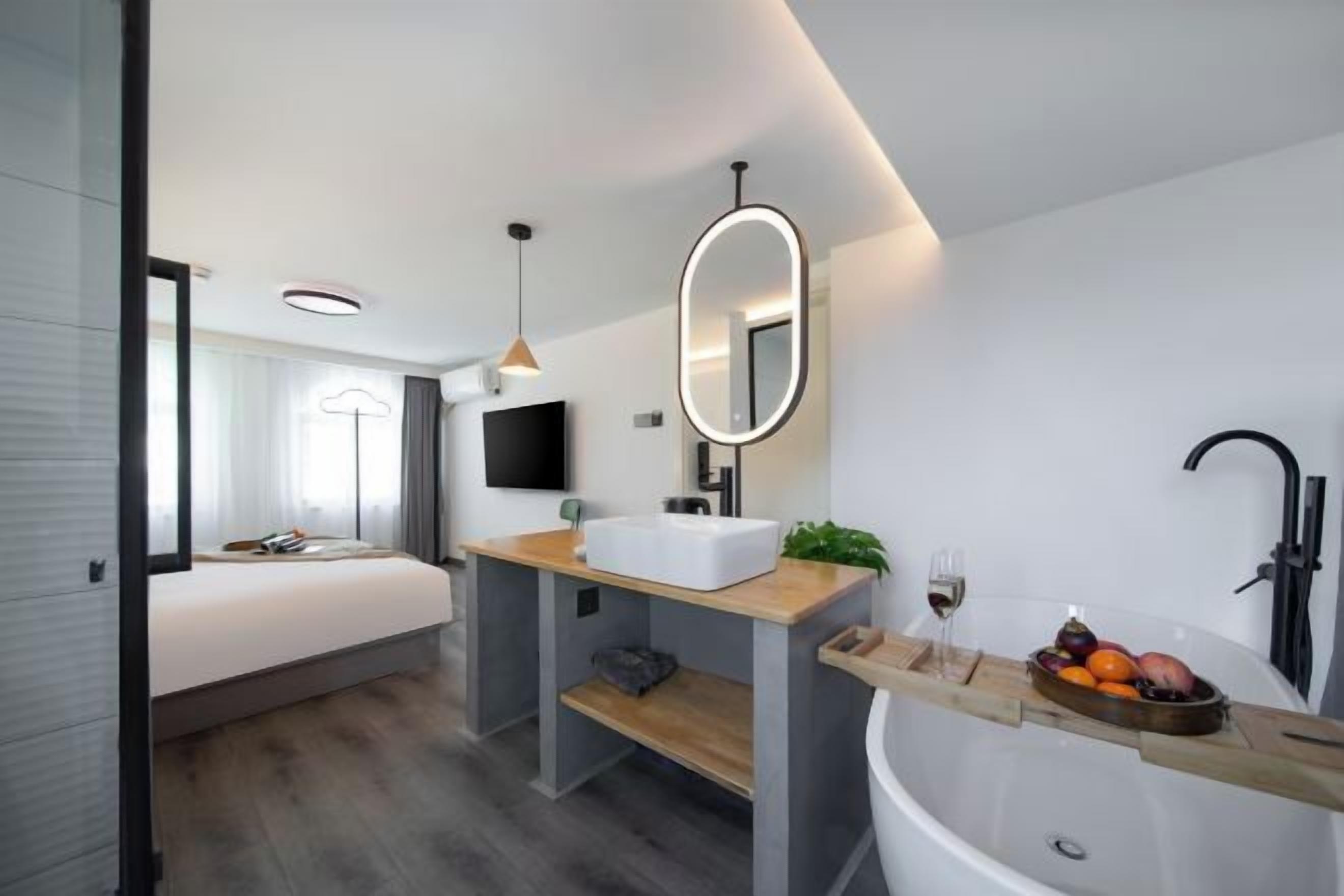 A modern hotel room with a large bed, a wooden vanity, a bathtub, and a small seating area.