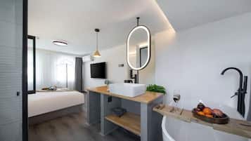 A modern hotel room with a large bed, a wooden vanity, a bathtub, and a small seating area.