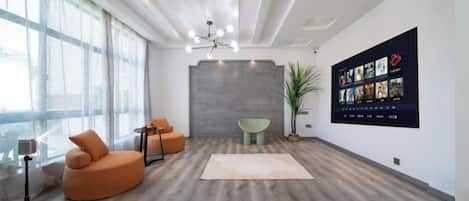 A modern interior with a wooden floor, a large window, a seating area with a sofa and armchairs, a wall-mounted display, and a ceiling light fixture.