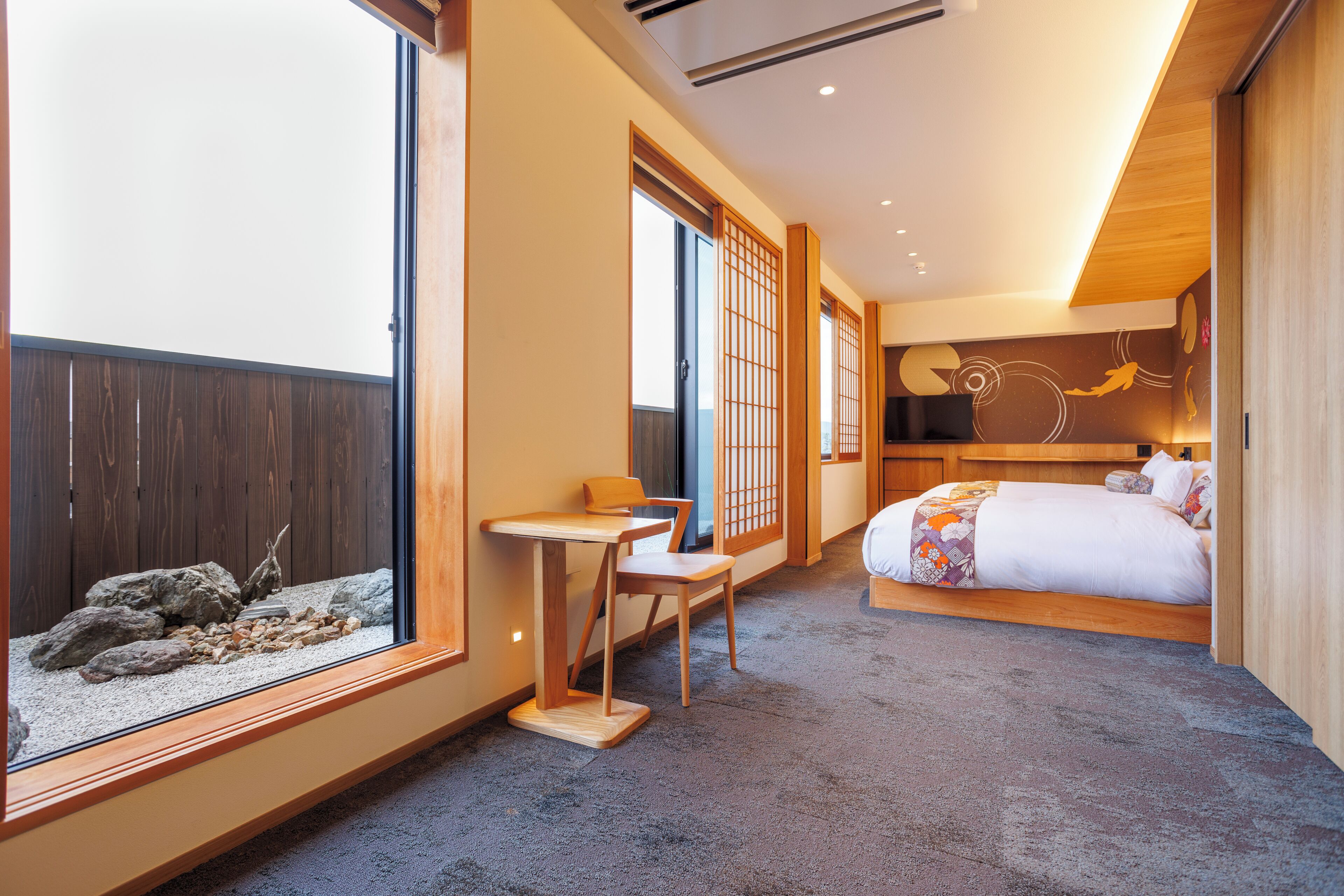 A hotel room with a bed, a small table, and a chair. There is a window with a view of a wooden fence and rocks.