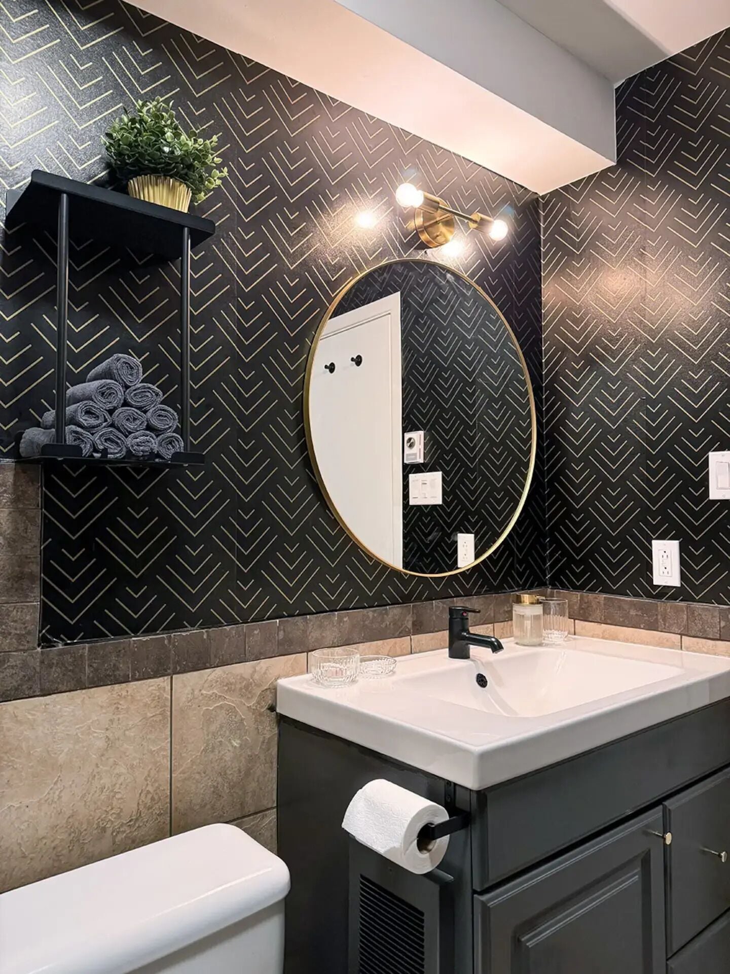 Een moderne badkamer met een muur in zwart-wit chevrons, een ronde spiegel, een witte wastafel en een zwart kastje.