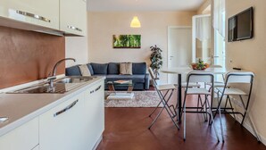 A compact living space with a kitchenette, a sofa, a dining area with chairs, and a TV mounted on the wall. - Nice apartment for 4 people with WIFI, TV and balcony (Osteno)