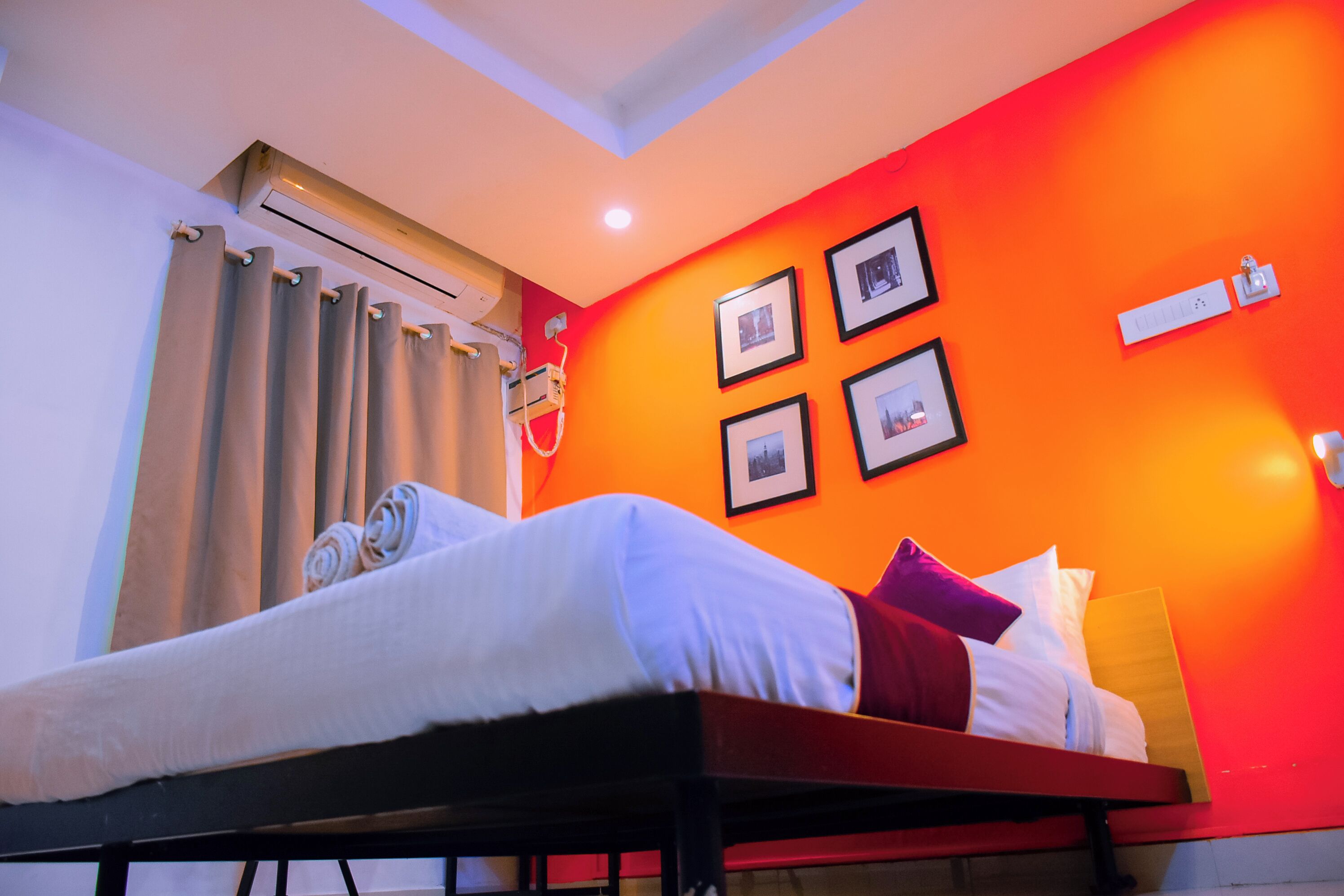 A hotel room with a bed, a window with curtains, an air conditioning unit, and framed pictures on the wall.