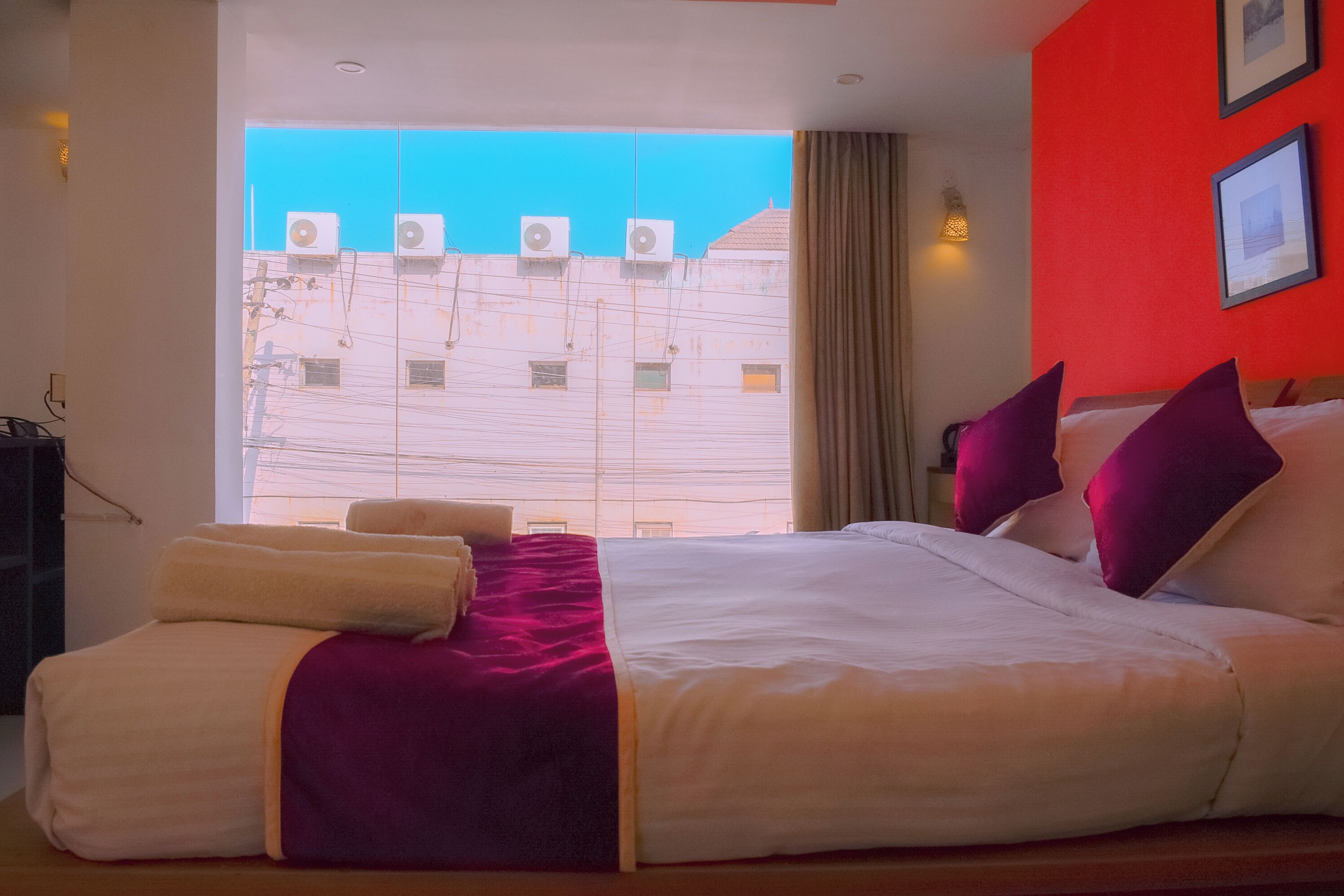 Una habitación de hotel con una cama grande, vista del paisaje urbano a través de una ventana y una pared roja.