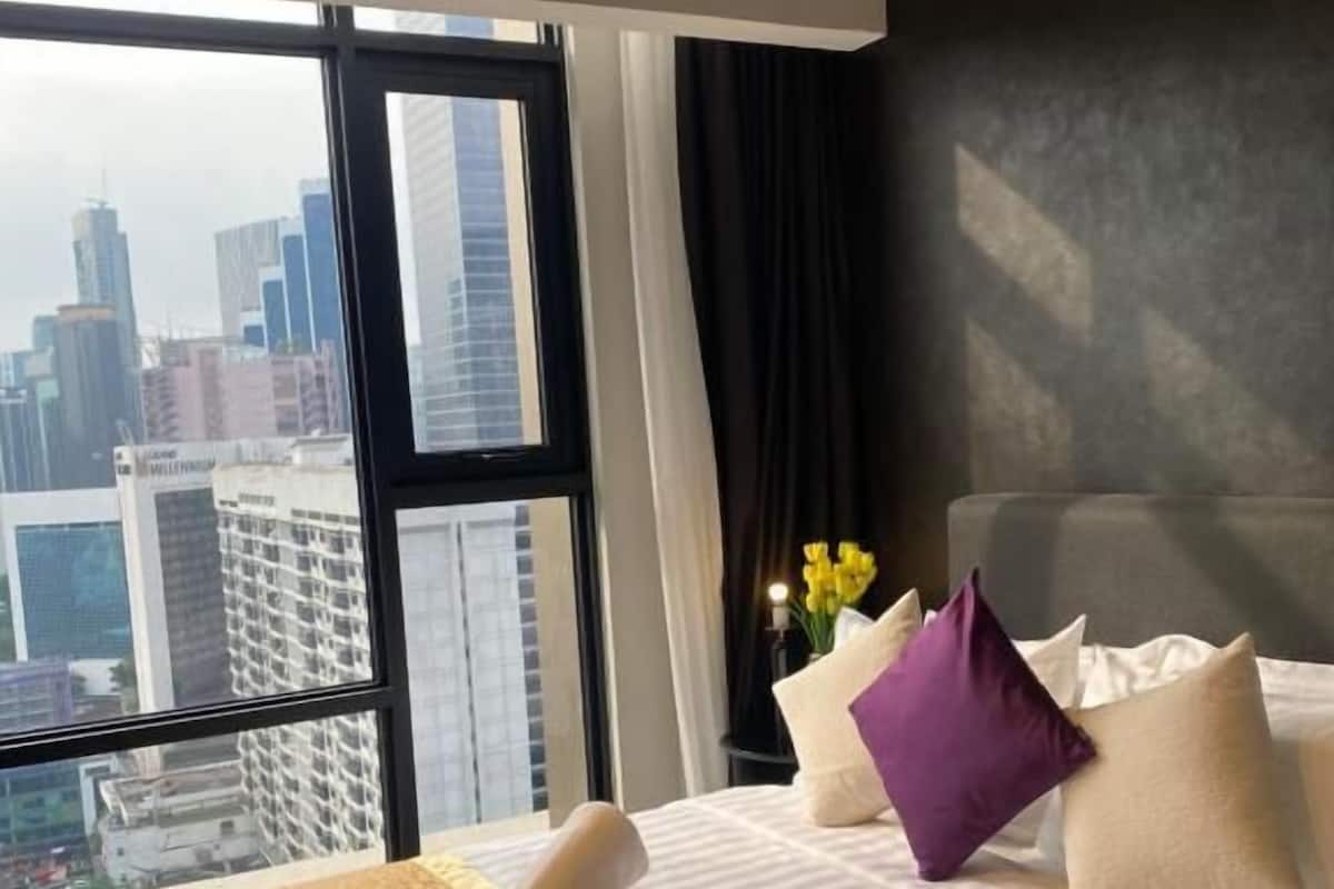 Ein Hotelzimmer mit großem Fenster und Blick auf die Stadt, einem ordentlich bezogenen Bett mit gemustertem Überwurf und einer Vase mit Blumen auf dem Nachttisch.