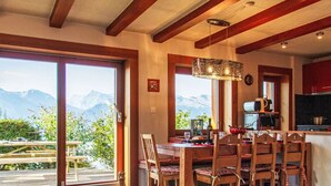 Un coin repas avec une table dressée pour deux personnes, une bouteille de vin et une vue sur les montagnes par de grandes fenêtres.