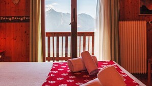 Une chambre d’hôtel avec un intérieur en bois, un lit recouvert d’une courtepointe à motifs floraux rouges, deux serviettes enroulées, une vue sur les montagnes à travers une porte vitrée et un radiateur fixé au mur.