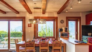 Un coin repas avec une table et des chaises en bois, une cuisine équipée d’un four à micro-ondes et d’une cuisinière, et de grandes fenêtres offrant une vue sur la verdure et les montagnes.