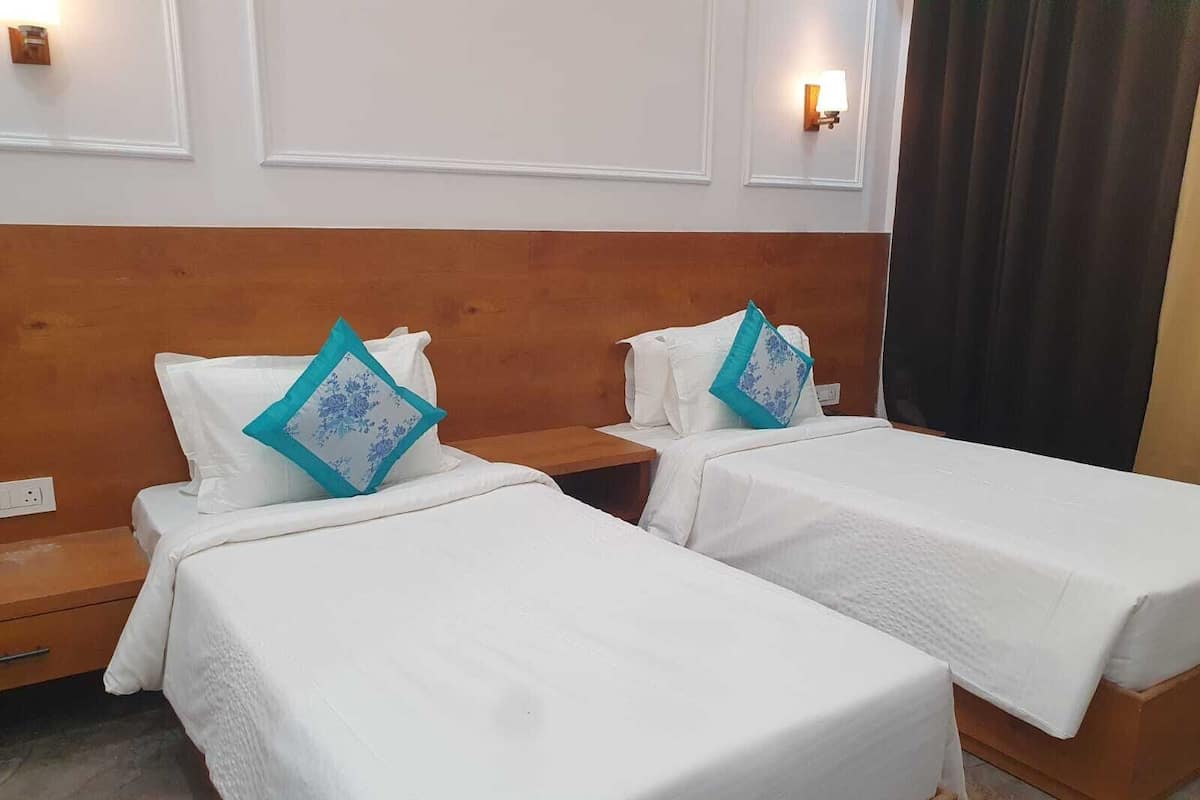 A hotel room with two single beds, each with a white bedspread and a blue floral pillow. There are wooden headboards and nightstands, and wall-mounted light fixtures.