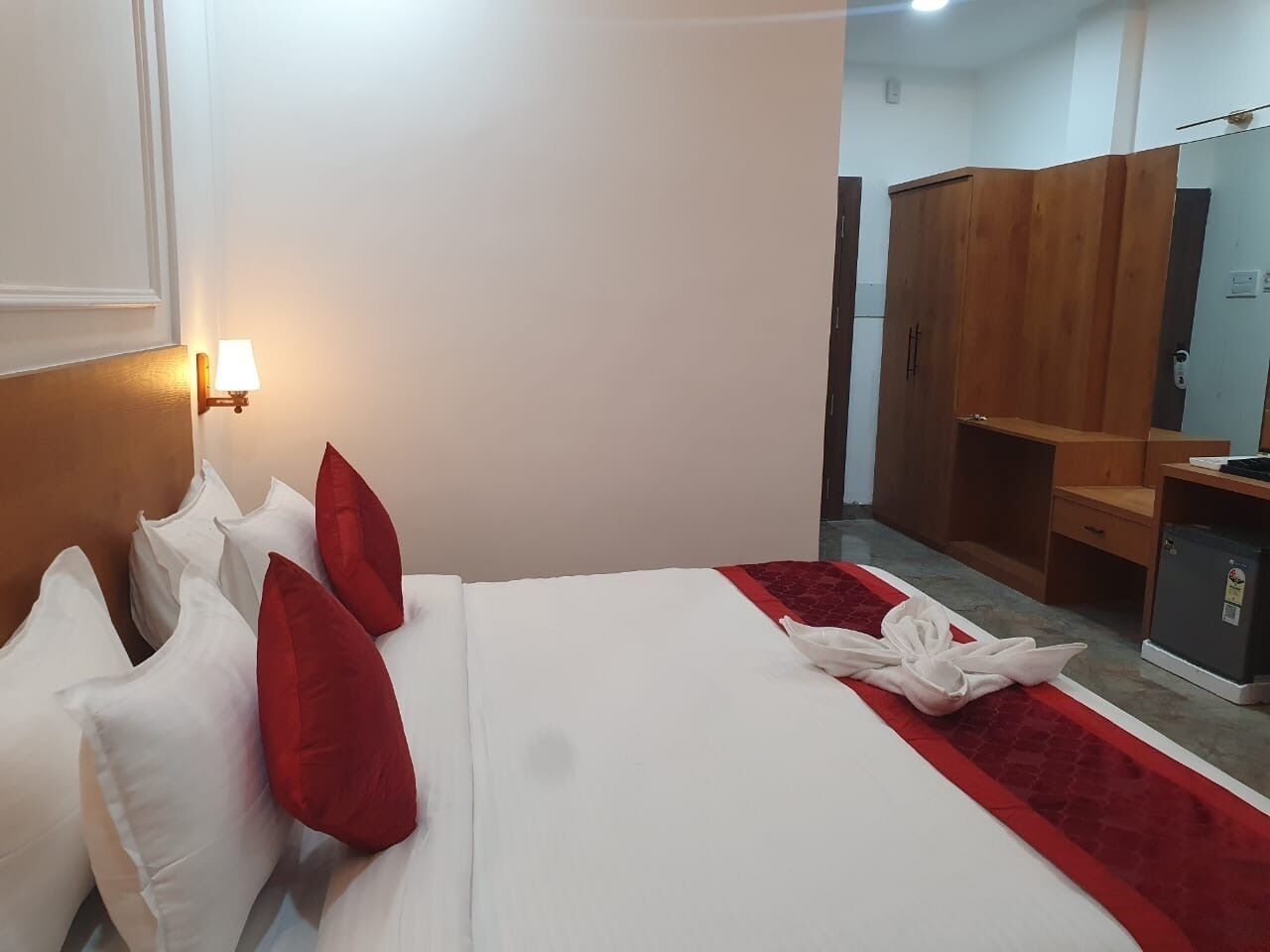 A hotel room with a neatly made bed, a red and white bedspread, and a white towel on the floor. There is a wooden wardrobe and a small refrigerator in the room.