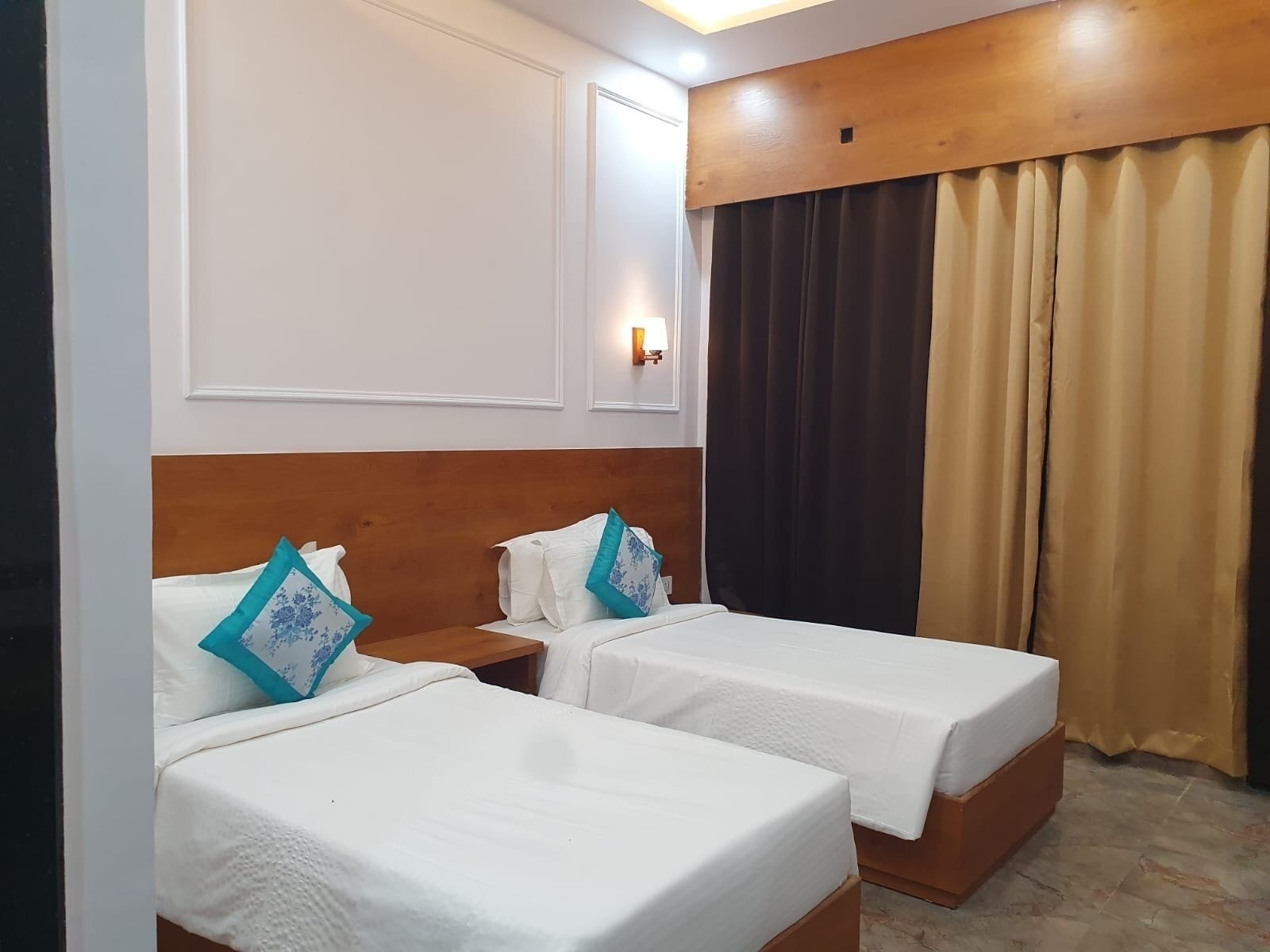 Habitación de hotel con dos camas individuales, cabecera de madera, mesita de noche, lámpara fijada a la pared y ventana con cortinas.