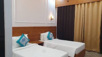 Habitación de hotel con dos camas individuales, cabecera de madera, mesita de noche, lámpara fijada a la pared y ventana con cortinas.