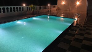 Een zwembad bij nacht met helder water, omringd door een zwart-wit schaakbordvloer en een witte balustrade.