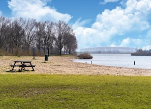 A park with a picnic table and a bench, a sandy area, and a body of water with a bridge in the background. - Bungalow in Zuytland With Private Fishing Jetty (Simonshaven)