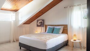A bedroom with a sloped ceiling, a bed with a wooden headboard, a bedside lamp, and a woven rug on the floor. - Résidence tamarin - nature lovers - sea and mountain views (LA RIVIERE)