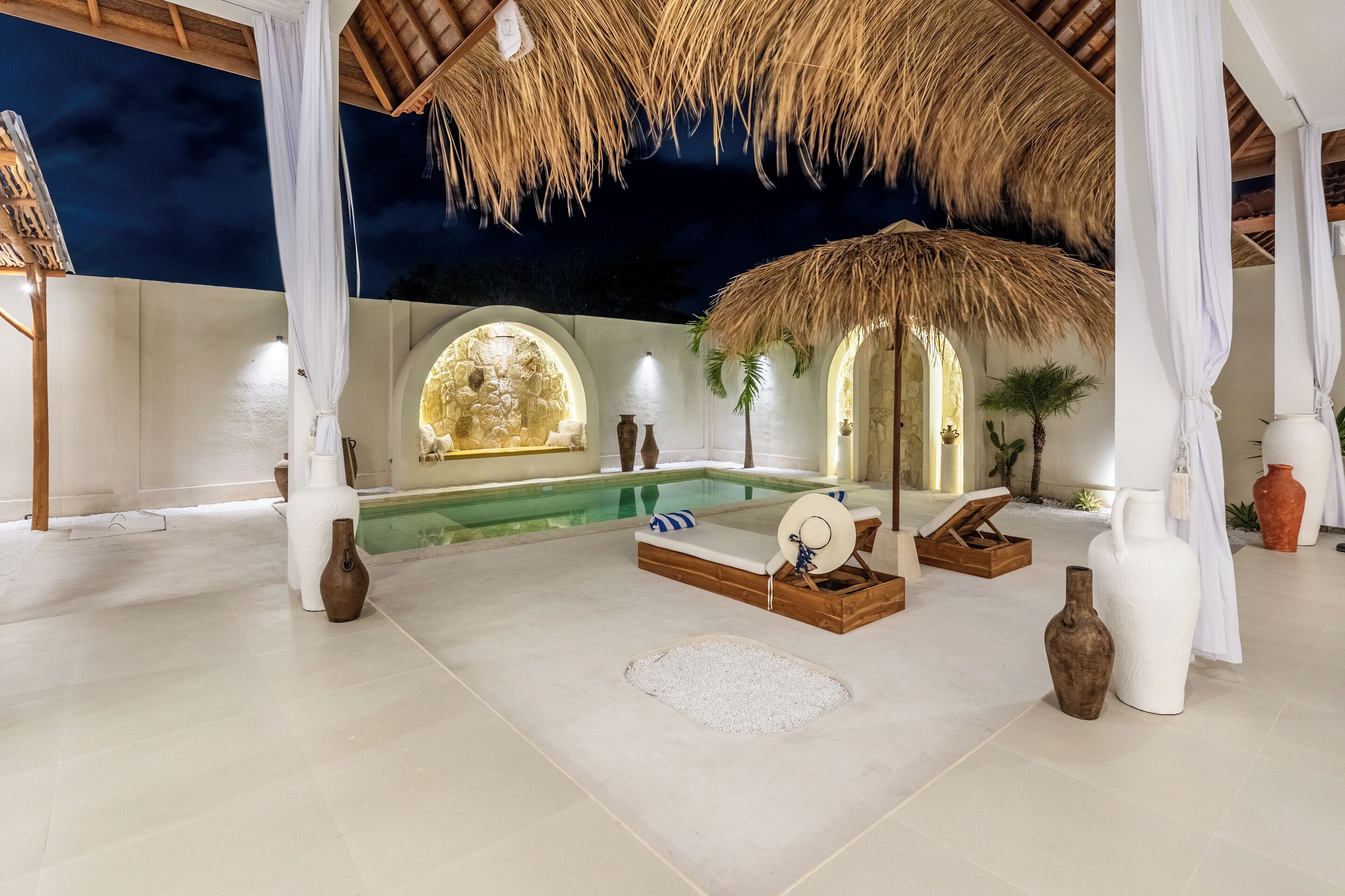 A luxurious indoor pool area with a thatched roof, white columns, and a stone wall backdrop.