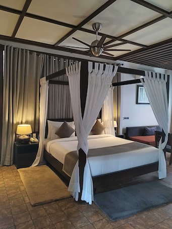A four-poster bed with a ceiling fan, a bedside table with a lamp, and a view of a cityscape through a window.. Pleasant Tree Hotels
