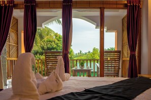A hotel room with a bed, a table with a plant, and a balcony with wooden railings and a view of greenery.