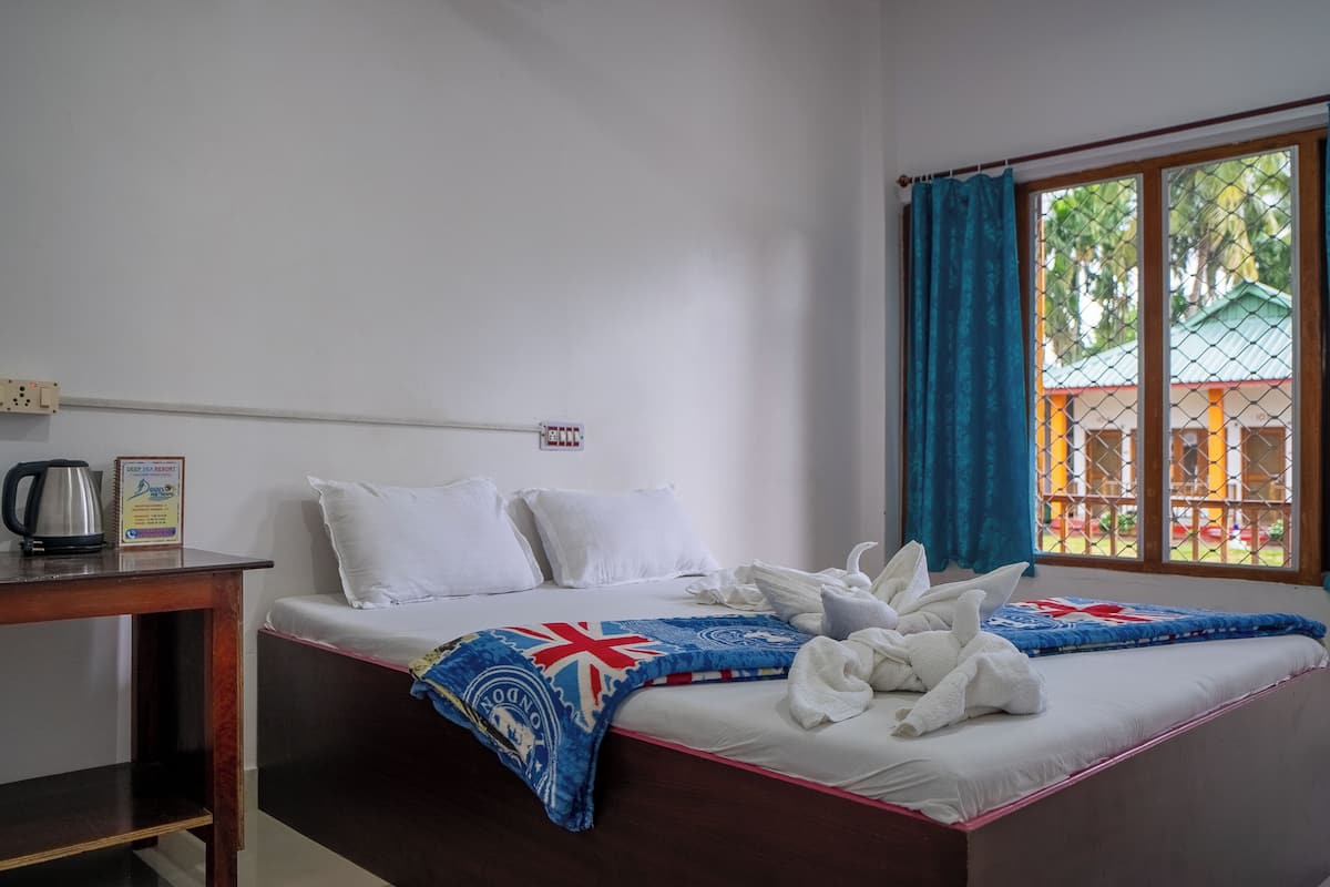A neatly made bed with white linens and a blue blanket with a Union Jack design, a wooden nightstand with a kettle, and a window with blue curtains.