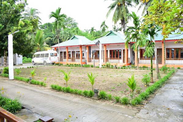A row of single-story buildings with green roofs, surrounded by a landscaped garden with small palm trees and shrubs.