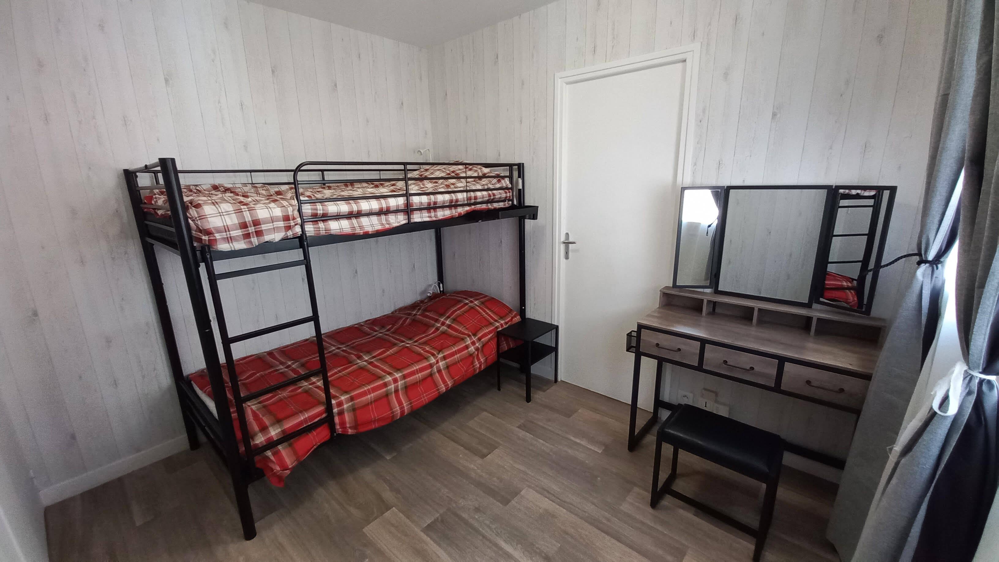 Ein Stockbett mit Karo-Bettwäsche, ein Schreibtisch mit Schubladen, ein Spiegel und eine Tür.