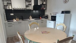 Eine kleine Küche mit weißen Schränken, einer schwarzen Dunstabzugshaube und einem weißen Kühlschrank. In der Mitte steht ein Esstisch mit vier Stühlen.