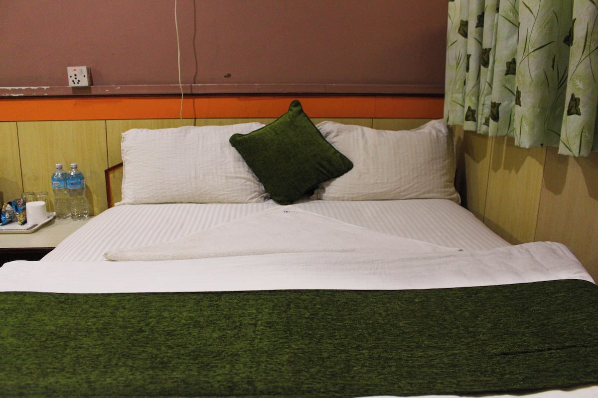 A hotel room with a bed, pillows, a green blanket, a tray with toiletries, and a wall socket.