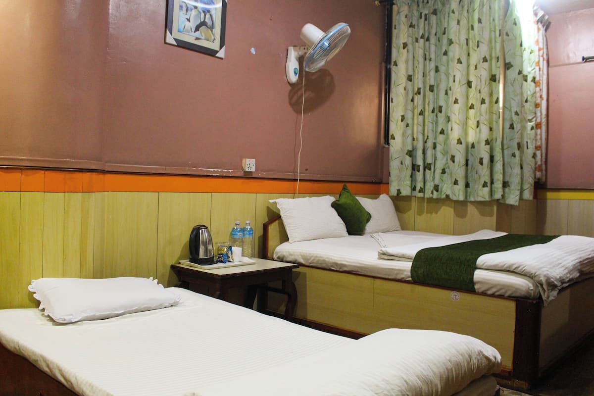 A hotel room with two single beds, a small table with a kettle and bottles, a wall-mounted fan, and a window with patterned curtains.