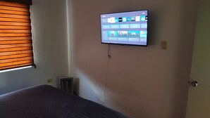 A room with a bed, a wall-mounted TV displaying various channels, and a window with blinds.