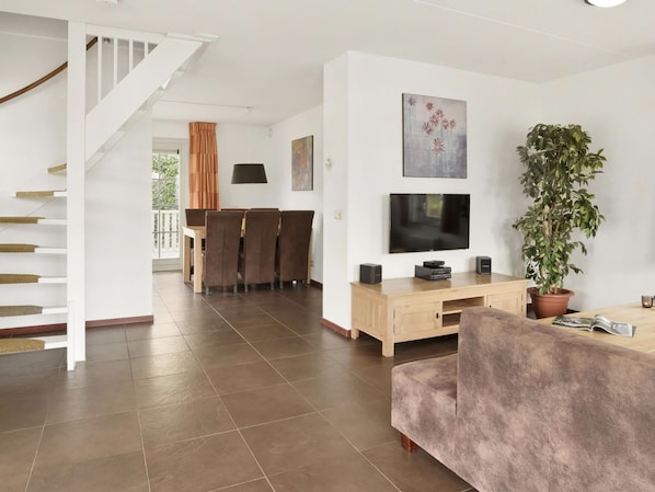 Una sala moderna con una escalera, un comedor, un televisor de pantalla plana, una planta grande en maceta y un mueble de entretenimiento de madera.